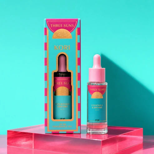 Three Suns Kori fragrance oil bottle with dropper next to its colorful packaging box, displayed on a pink acrylic pedestal against a turquoise background.