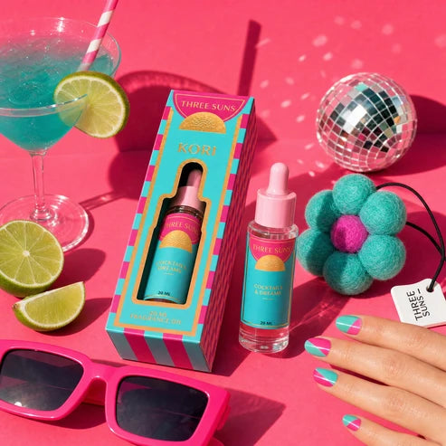 Three Suns Kori fragrance oil shown in its striped box and as an open dropper bottle, styled on a hot pink background with tropical drinks, sunglasses, a disco ball, and a hand with matching pink and teal nail polish.