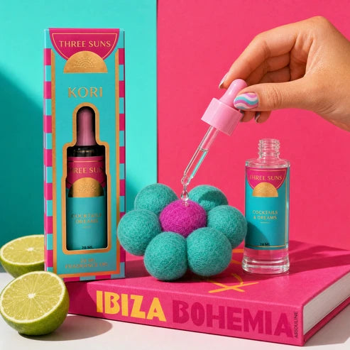 Three Suns Kori fragrance oil set shown with its box, a glass dropper bottle, and a small matching bottle on a bright pink and turquoise set. A hand is holding the dropper above a flower-shaped object, with lime halves and an Ibiza Bohemia book in the scene.