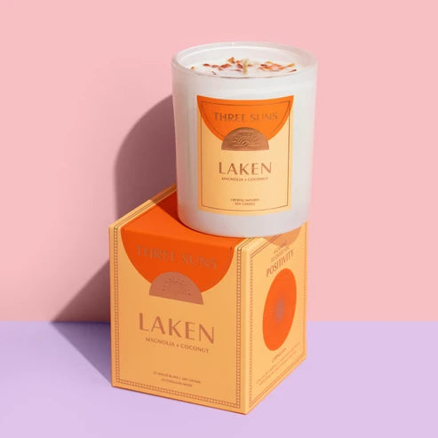 A Three Suns Laken scented candle in a white glass jar sits on top of its matching orange and beige packaging box against a split pink and lavender background.