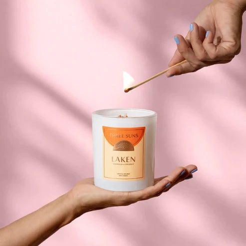 A white candle jar labeled 'Three Suns Laken' is held in a hand while another hand ignites the candle with a wooden match against a soft pink background.