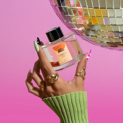 A hand with green, pink, and white nail art holds a clear glass perfume bottle labeled &