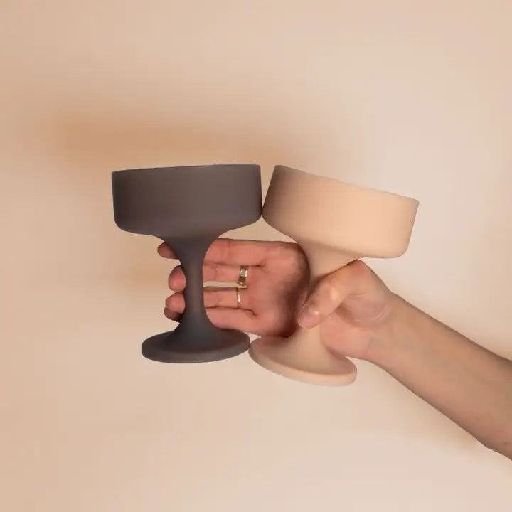 Two matte silicone cocktail glasses in dark gray and light beige held side by side in two hands against a warm beige background.