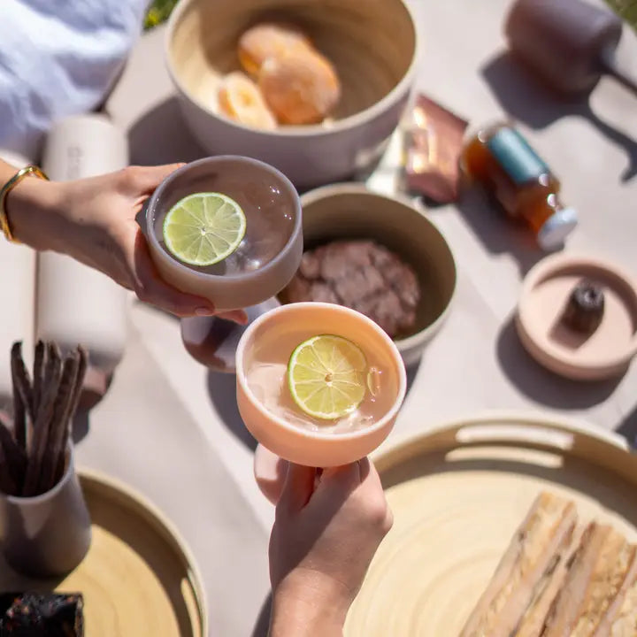 Two hands holding pastel-colored silicone cocktail glasses with lime slices, set in a bright, sunlit outdoor dining environment with wooden trays and bowls.