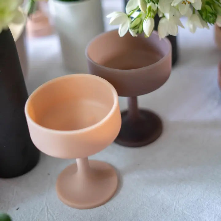 Two matte silicone cocktail glasses in dusty peach and taupe brown, styled on a light fabric surface with blurred white flowers and dark vases in the background.