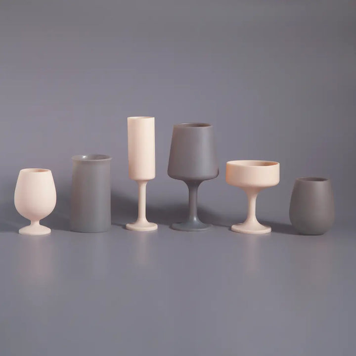 Six silicone drinkware pieces in neutral shades of cream and gray, arranged in a row on a matching gray surface against a gray background.