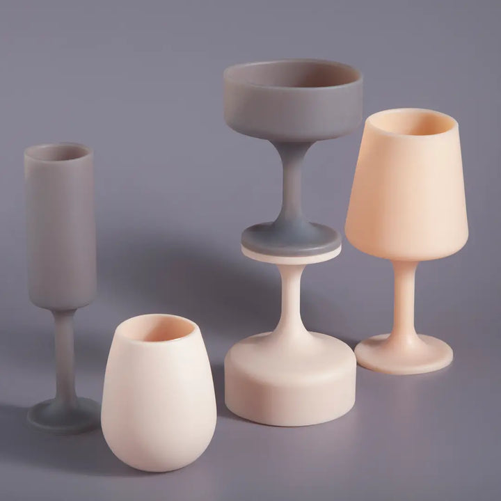 Five silicone drinkware pieces in neutral tones of gray, cream, and pale peach, arranged on a smooth gray surface against a matching gray background.
