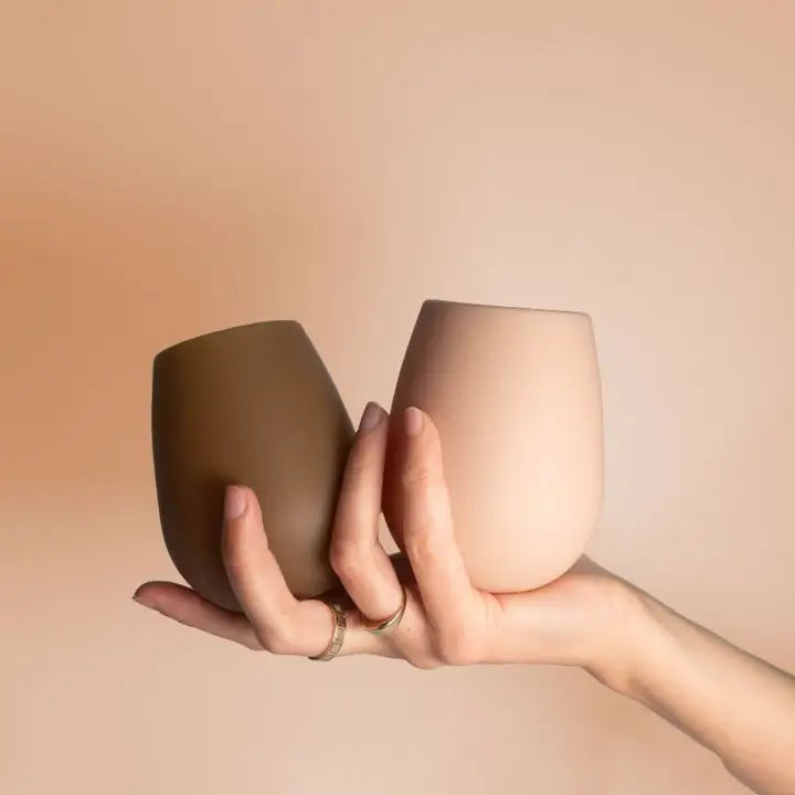 Two silicone stemless wine glasses in earthy tones, one dark brown and one light beige, held in a person&