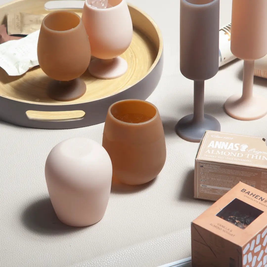 A styled product shot of silicone drinkware in soft neutral tones, including two small stemless cups, two wine glasses, and two rounded vessels, arranged on a light surface with a round tray and product boxes nearby.