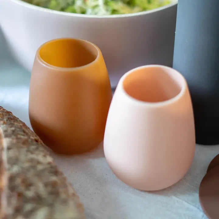 Two silicone unbreakable glass wine tumblers, one in warm amber-brown and one in pale blush pink, shown on a tabletop with food props in the background.