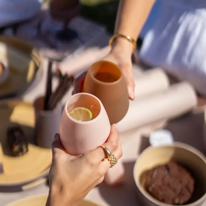 Two hands clinking pastel silicone drinking glasses in a bright outdoor setting. One glass is light pink with a lime slice, and the other is a warm terracotta brown.