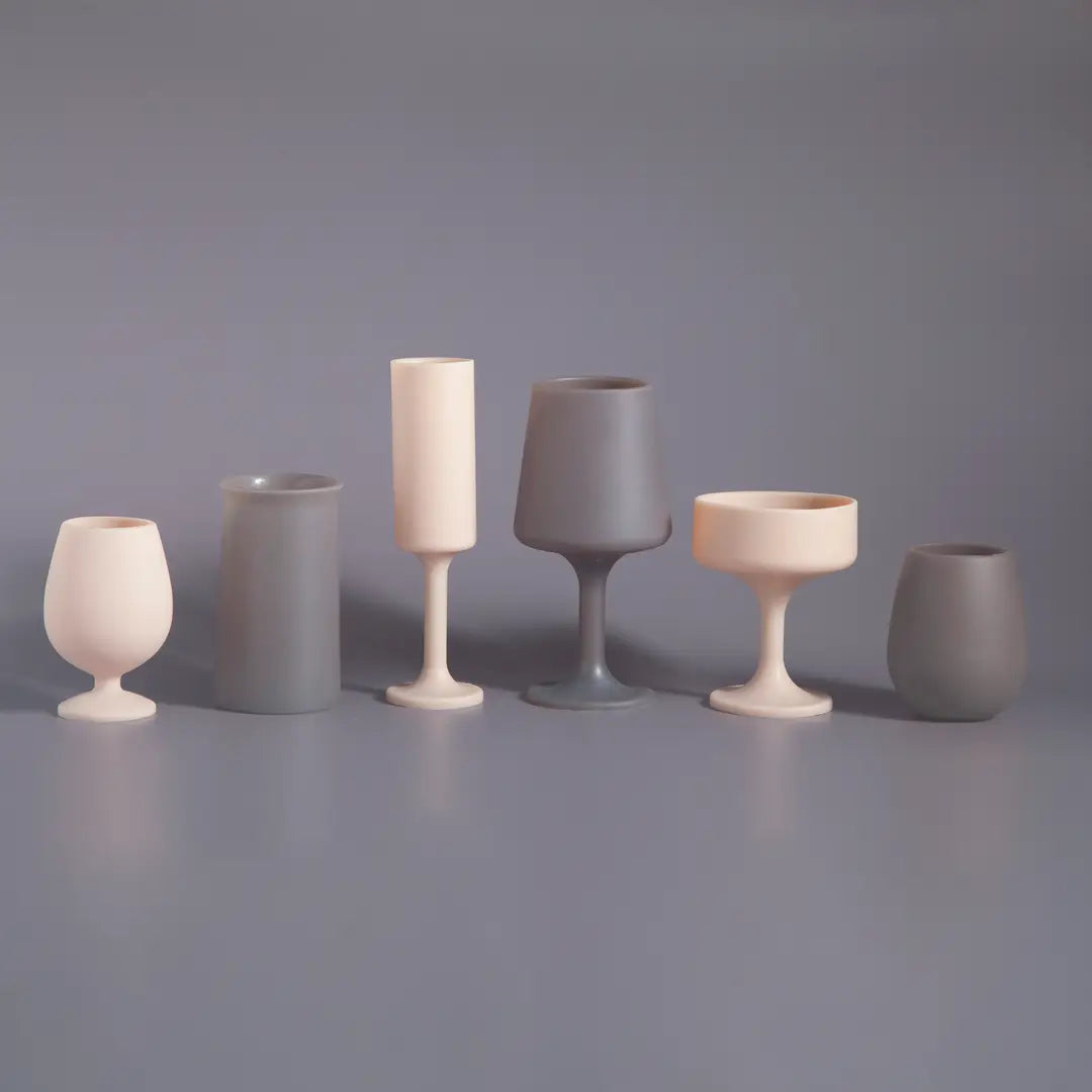 Six silicone unbreakable glasses in neutral shades of cream and gray, arranged in a row on a matching gray surface against a gray background.