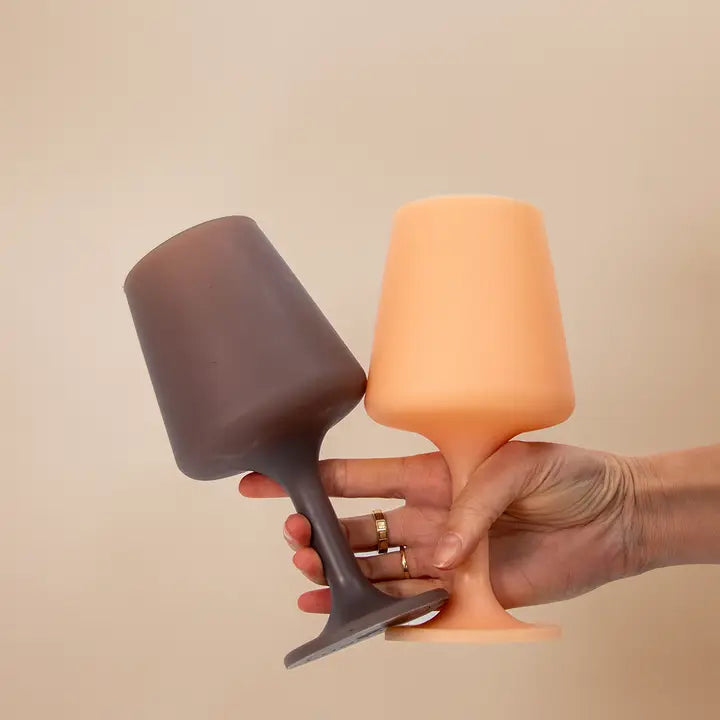 Two modern silicone wine glasses in matte dark brown and peachy orange, held by a hand against a plain beige background.