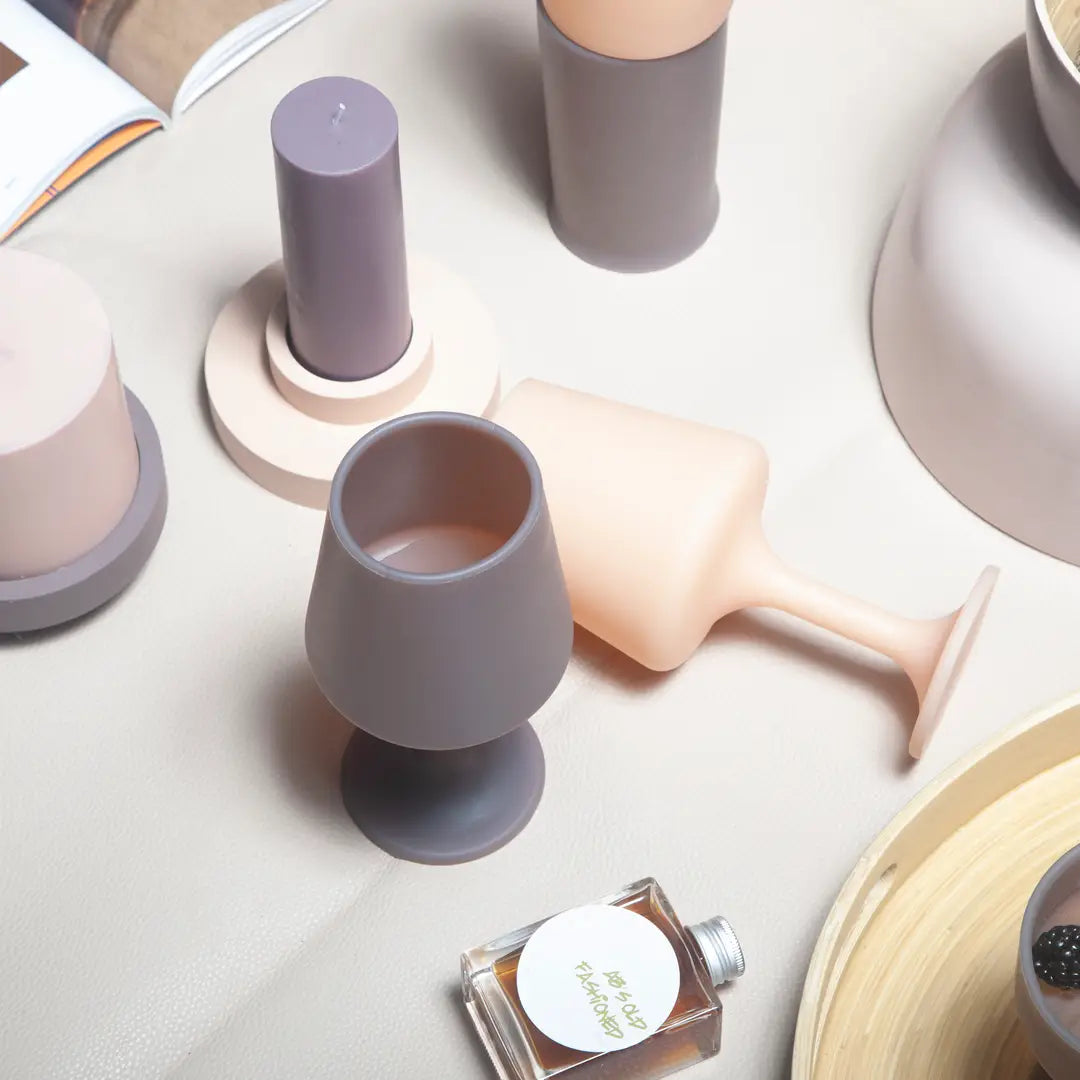 A styled flat-lay of matte home decor and fragrance items in muted pastel tones, including a gray goblet-shaped candle holder, a pale peach wine glass, matching candle bases, and a small perfume bottle on a light beige surface.