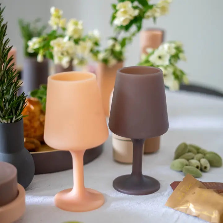 Two matte silicone wine glasses, one in a soft peach-orange color and the other in a dark taupe-brown, displayed on a white tablecloth with blurred floral and greenery props in the background.