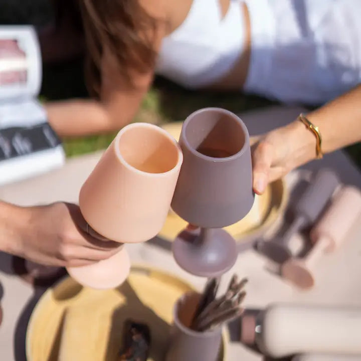Two silicone wine glasses, one in dusty pink and one in muted taupe, clinking together in an outdoor setting with a woman holding them.