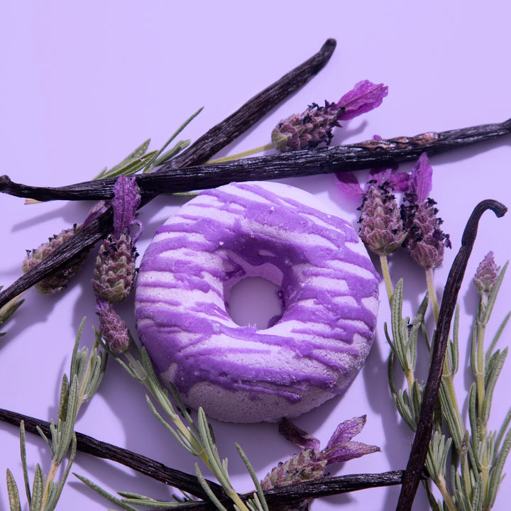 Purple and white donut-shaped bath bomb surrounded by lavender flowers and vanilla beans on a purple background