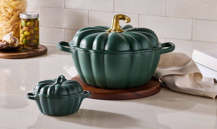 A dark green pumpkin-shaped lidded casserole dish with a gold-toned handle, shown on a kitchen counter beside a matching small ramekin, with pasta and a jar in the background.