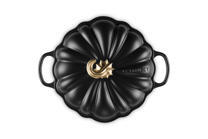 A black cast iron Dutch oven with a lid designed in a pumpkin shape, featuring a polished gold-toned metal central knob and the &