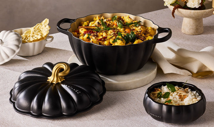 Black pumpkin-shaped ceramic casserole dish with a matching lid, styled on a beige tablecloth with food dishes and napkins around it.