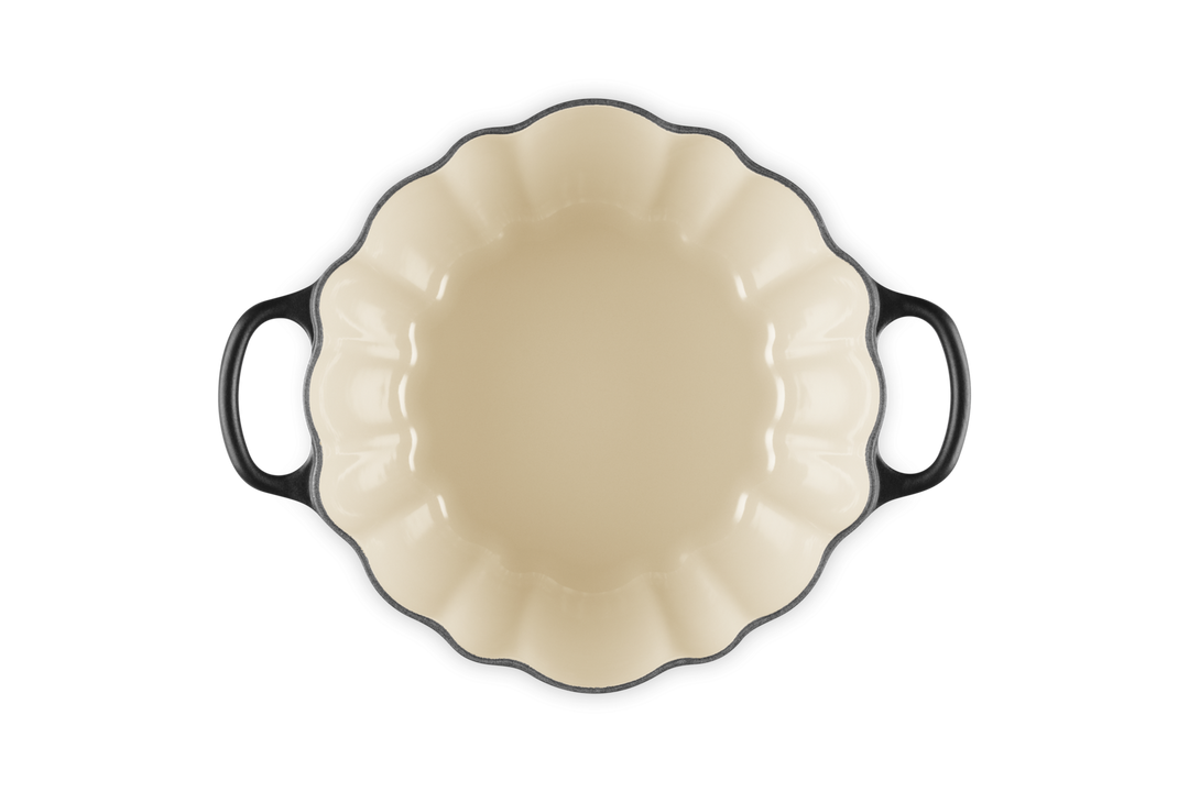 A baking dish with a cream-colored interior and black exterior, featuring a scalloped rim and two side handles, shown from a top-down perspective on a white background.