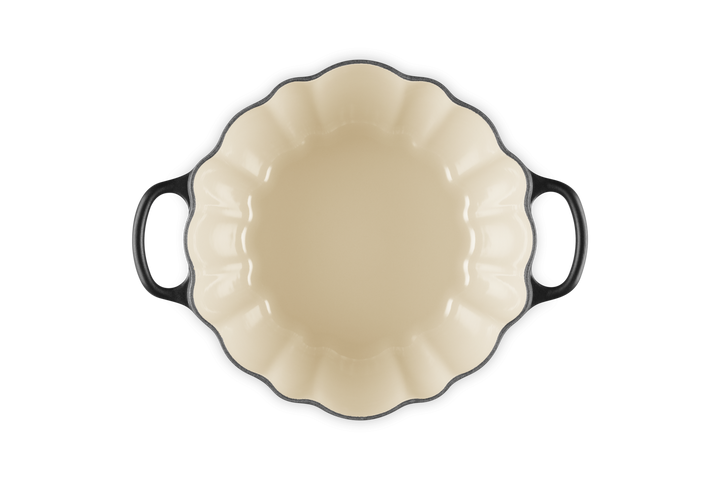 A baking dish with a cream-colored interior and black exterior, featuring a scalloped rim and two side handles, shown from a top-down perspective on a white background.