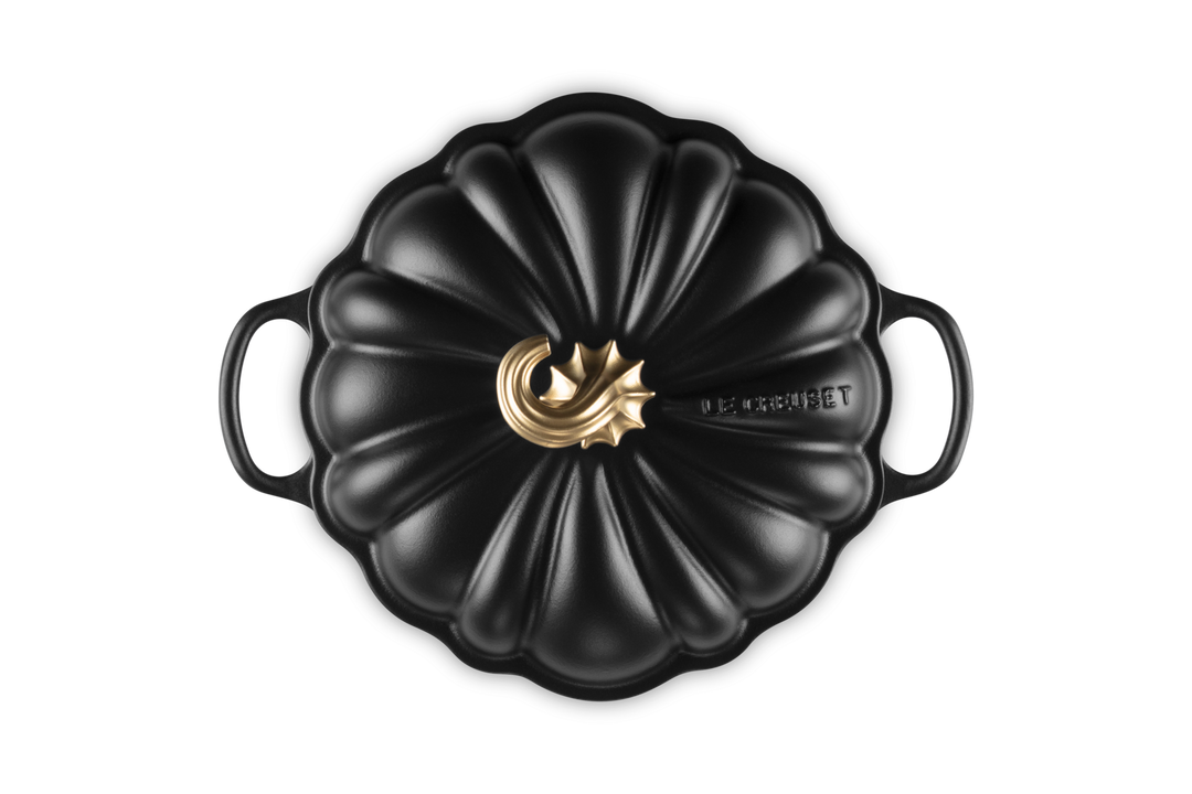 A black cast iron Dutch oven with a lid designed in a pumpkin shape, featuring a gold-toned metal central knob and the &