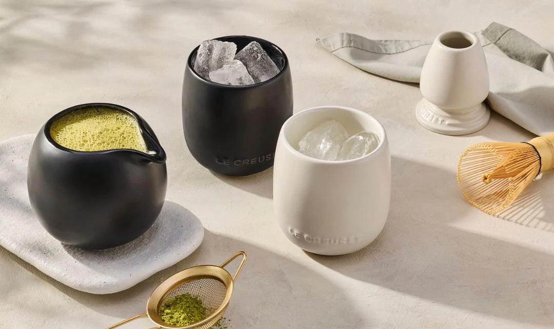 Le Creuset Serenity Collection - Jug with matcha in it, 2 ceramic tumblers with ice and a matcha whisk and stand on a beige surface. 