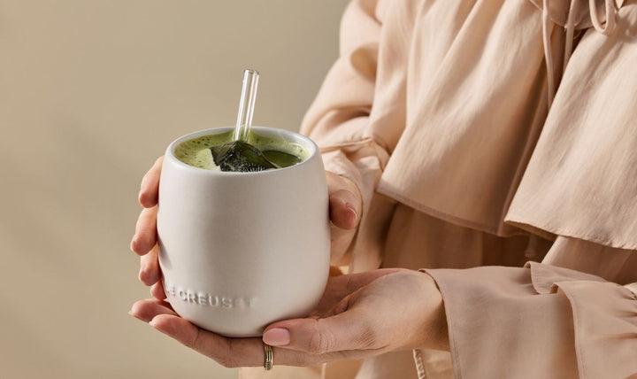 A person holding a brioche Le creuset ceramic tumbler with matcha in it with a glass straw