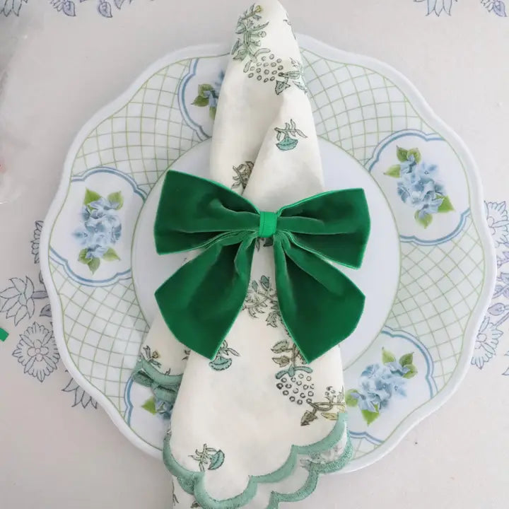 A folded white napkin with a green floral print and scalloped edge, tied with a large green velvet bow, presented on a white plate with a green grid pattern.