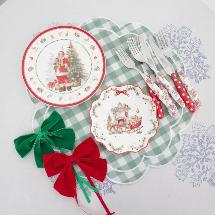 A styled holiday table setting featuring two scalloped Christmas dinner plates with red trim and festive illustrations, a set of silver forks with red and white patterned handles, and two large velvet bows in green and red on a white background with a green gingham placemat.