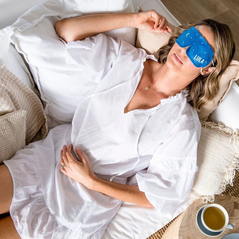 Woman wearing a blue sleep mask with 'Lula' branding, lying in bed with a cup of tea on a wooden table.