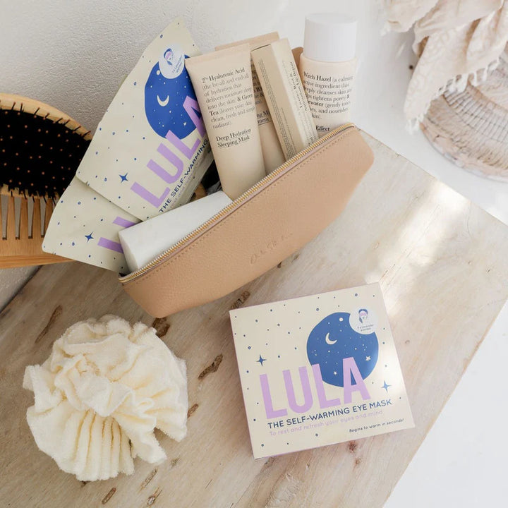 LULA self-warming eye mask packaging on a wooden surface with a hairbrush and books in the background.
