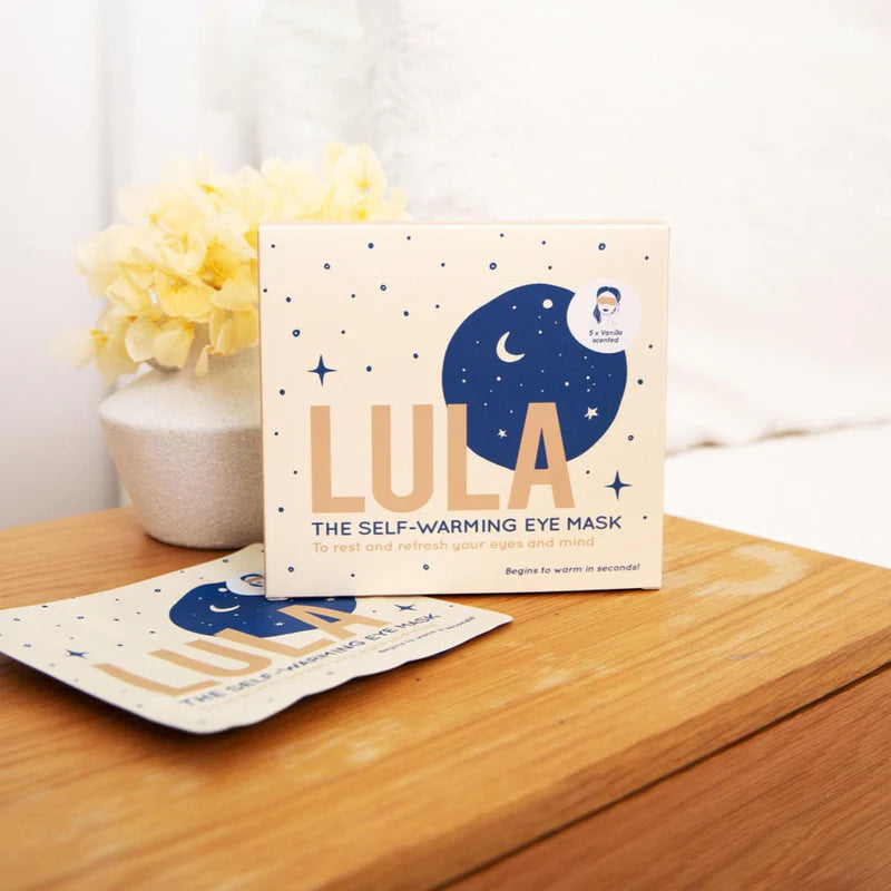 LULA self-warming eye mask packaging on a wooden surface with flowers in the background