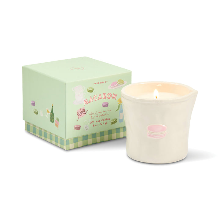 Candle with macaron design next to its packaging on a white background