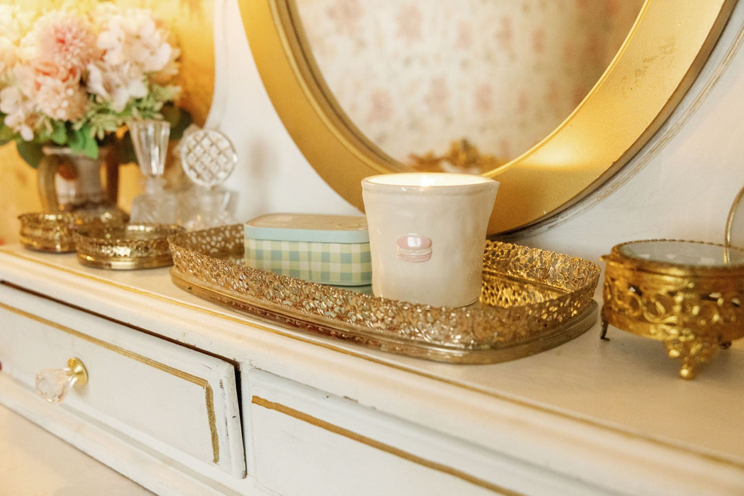 A close-up of a white ceramic candle with a pink bow on a decorative gold tray, styled on a vintage white dresser with gold trim next to a gold mirror.