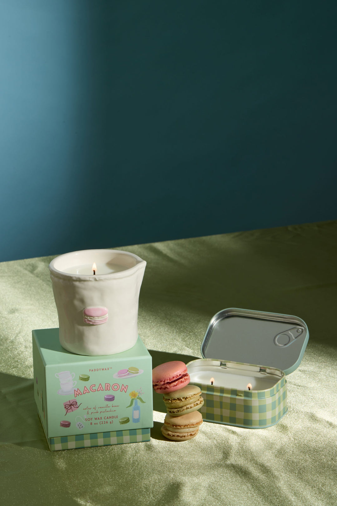 A styled product shot featuring a white ceramic candle in a small pink macaron-shaped holder, displayed on top of a mint green box. Next to it is an open round tin candle with a green-and-white checkered lid, alongside two macaron cookies.