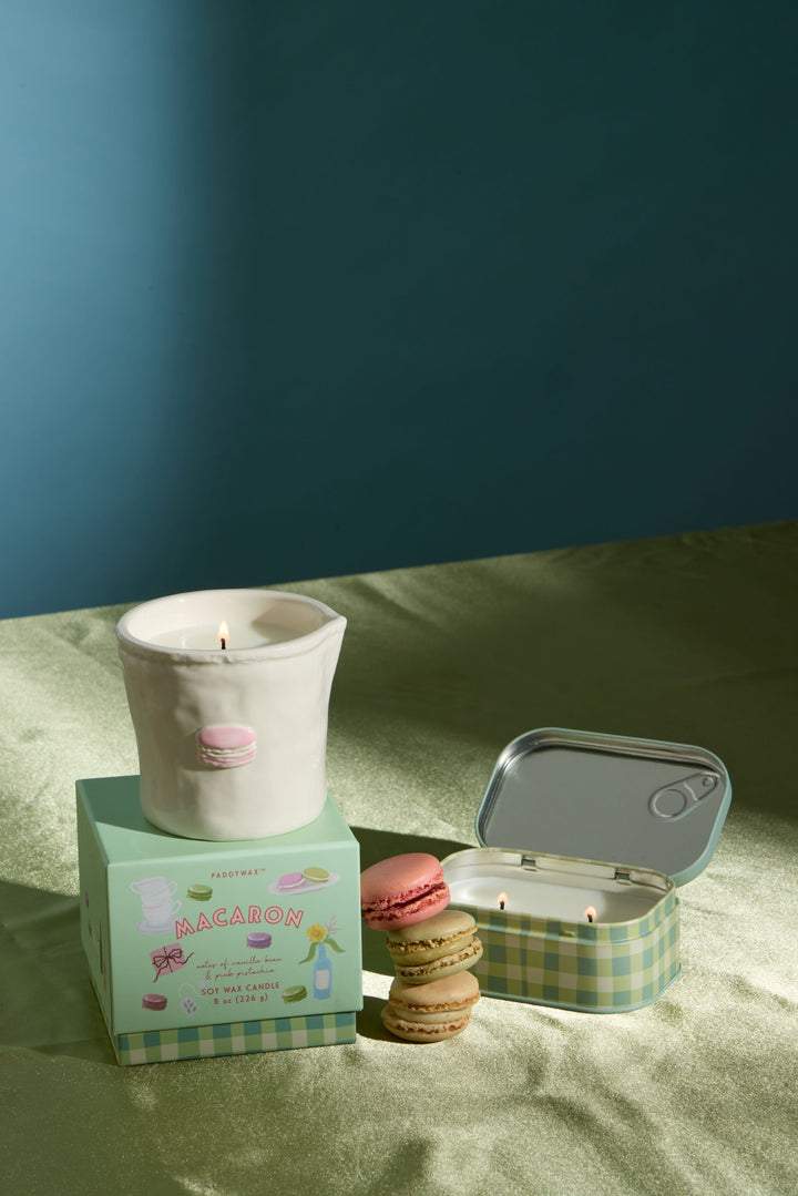 A styled product shot featuring a white ceramic candle in a small pink macaron-shaped holder, displayed on top of a mint green box. Next to it is an open round tin candle with a green-and-white checkered lid, alongside two macaron cookies.