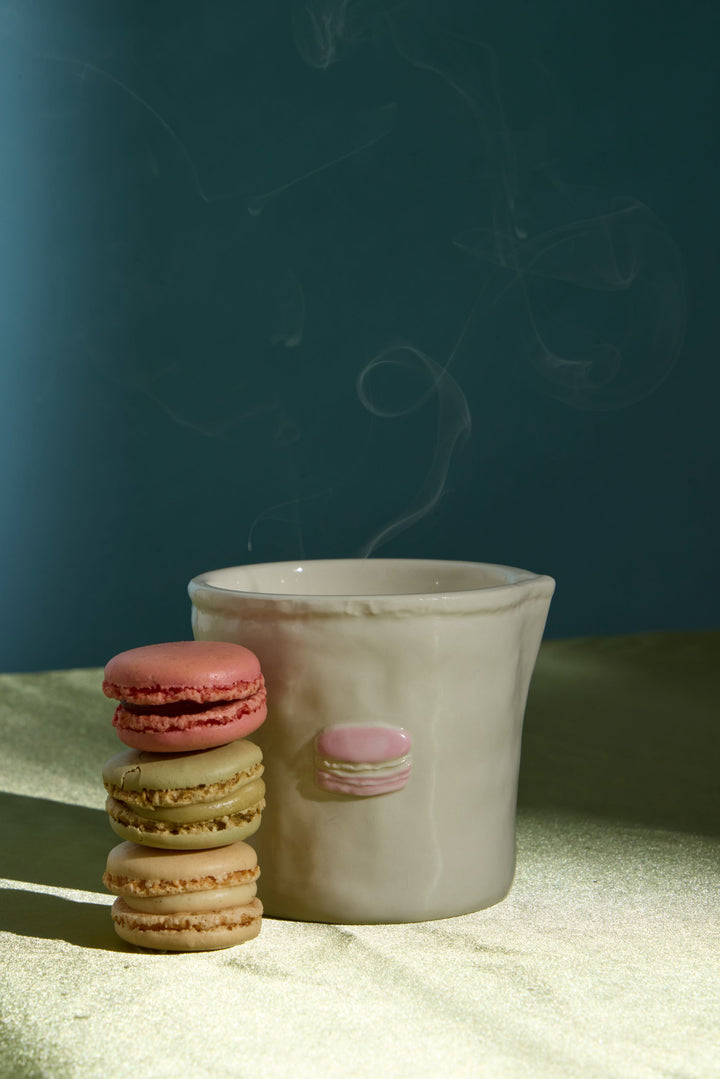 A small white ceramic cup with a pink macaronic accent sits on a textured cream surface next to a stack of three macarons (pink, green, and tan) against a dark teal background with soft steam rising.