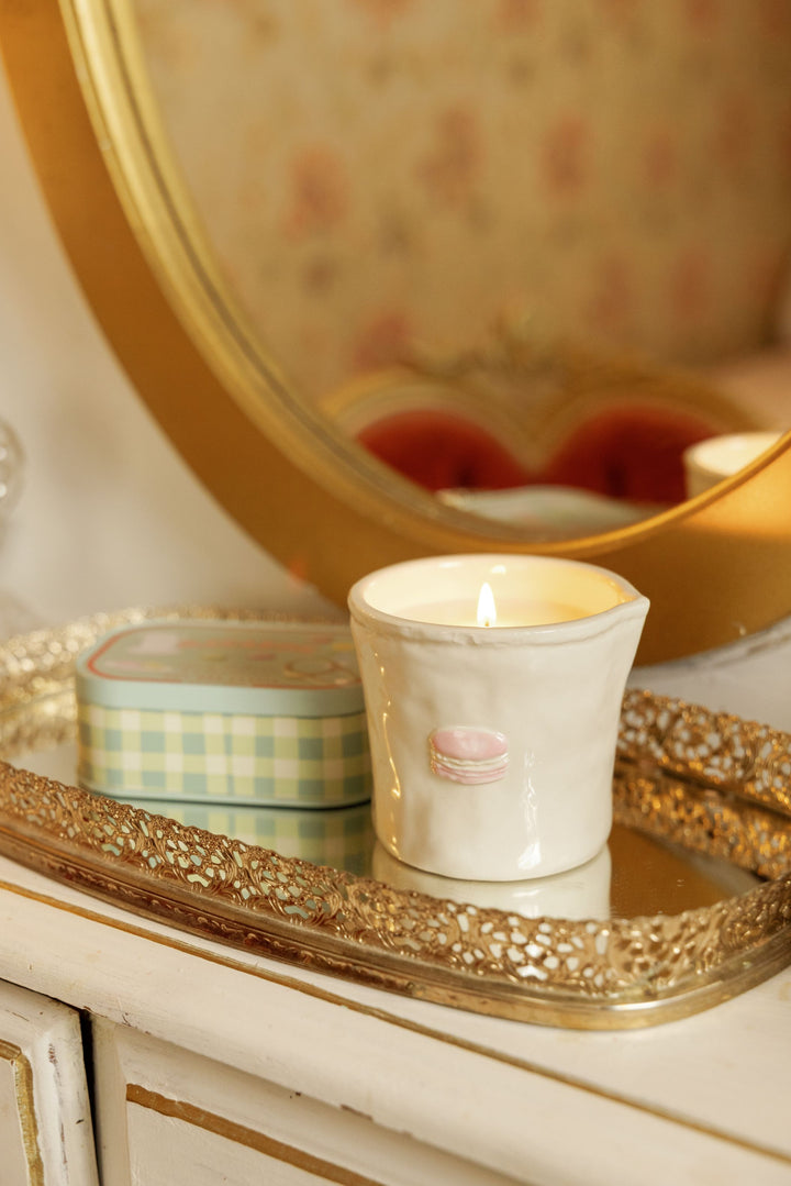 A white ceramic candle holder with a pink macaron graphic sits on a decorative gold tray next to a pastel green and white checkered tin, set against a vintage-style floral mirror.