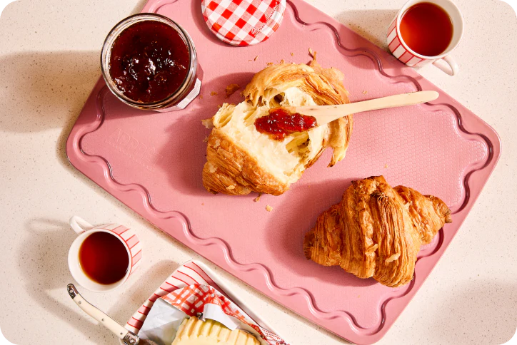 Breakfast scene with croissants, jam, and tea on a pink cutting board.