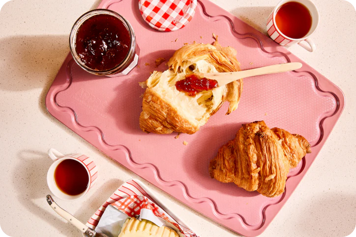 Breakfast scene with croissants, jam, and tea on a pink cutting board.
