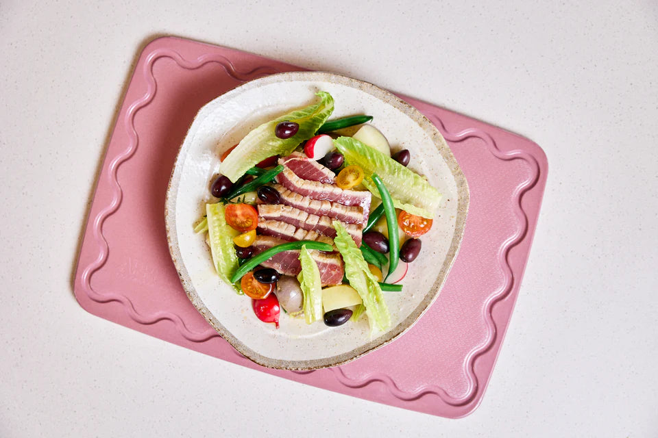 Salad with greens, tomatoes, and black olives on a white plate with a pink cutting board underneath.