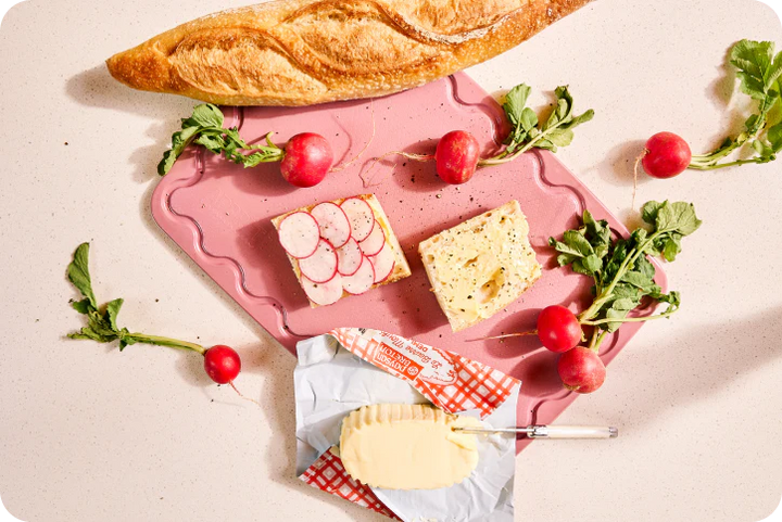 Pink cutting board with bread, radishes, and a spread on a light background