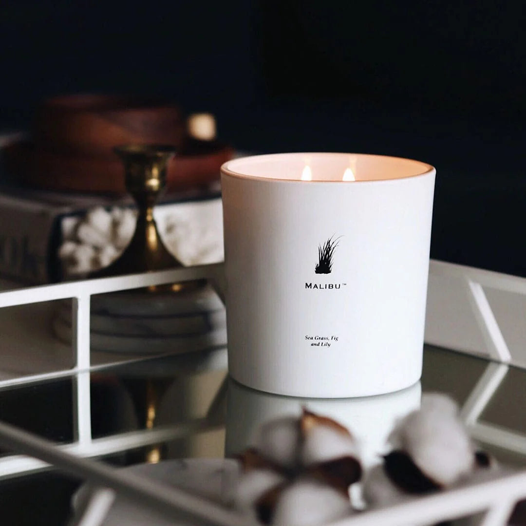 White candle with 'Malibu' branding on a reflective surface with dark background