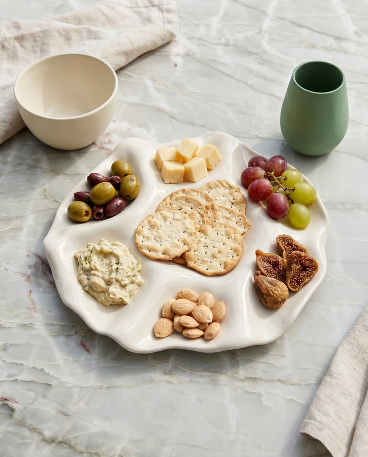 A white ceramic charcuterie board with five compartments, styled with crackers, cheese, olives, dip, nuts, and dried figs on a light marble surface, with a cream bowl and a green cup nearby.