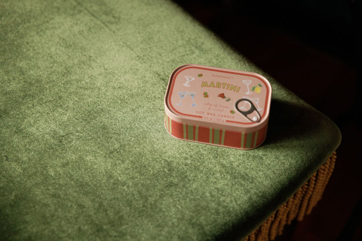 A small decorative tin candle labeled MARTINI sits on a textured green velvet surface in a dimly lit setting.