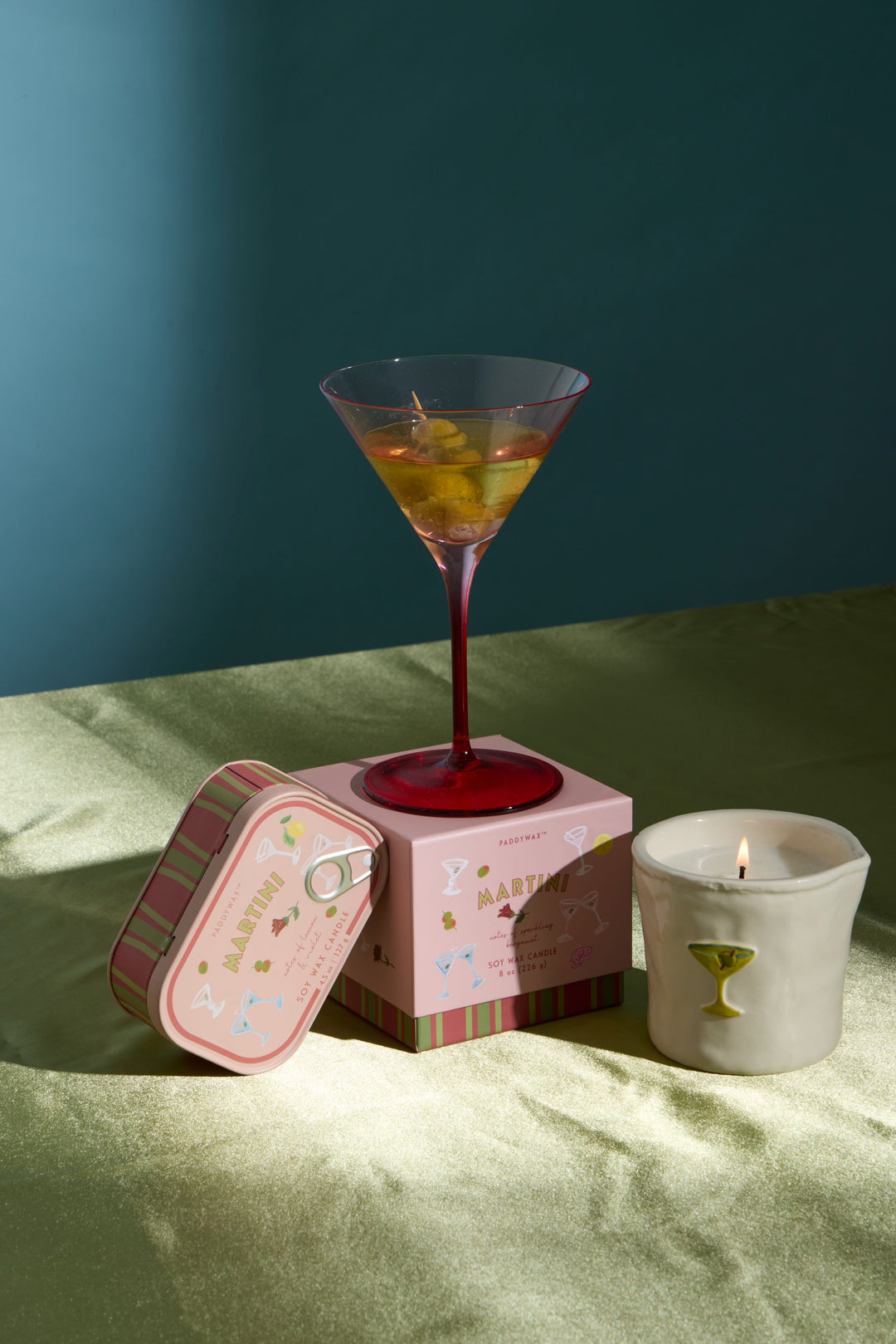 A styled product arrangement on a draped green fabric surface against a dark teal wall. A pink martini glass candle holder with a red stem and filled coupe glass sits on top of a pink box, next to a pink striped tin and a white candle with a green martini glass emblem.