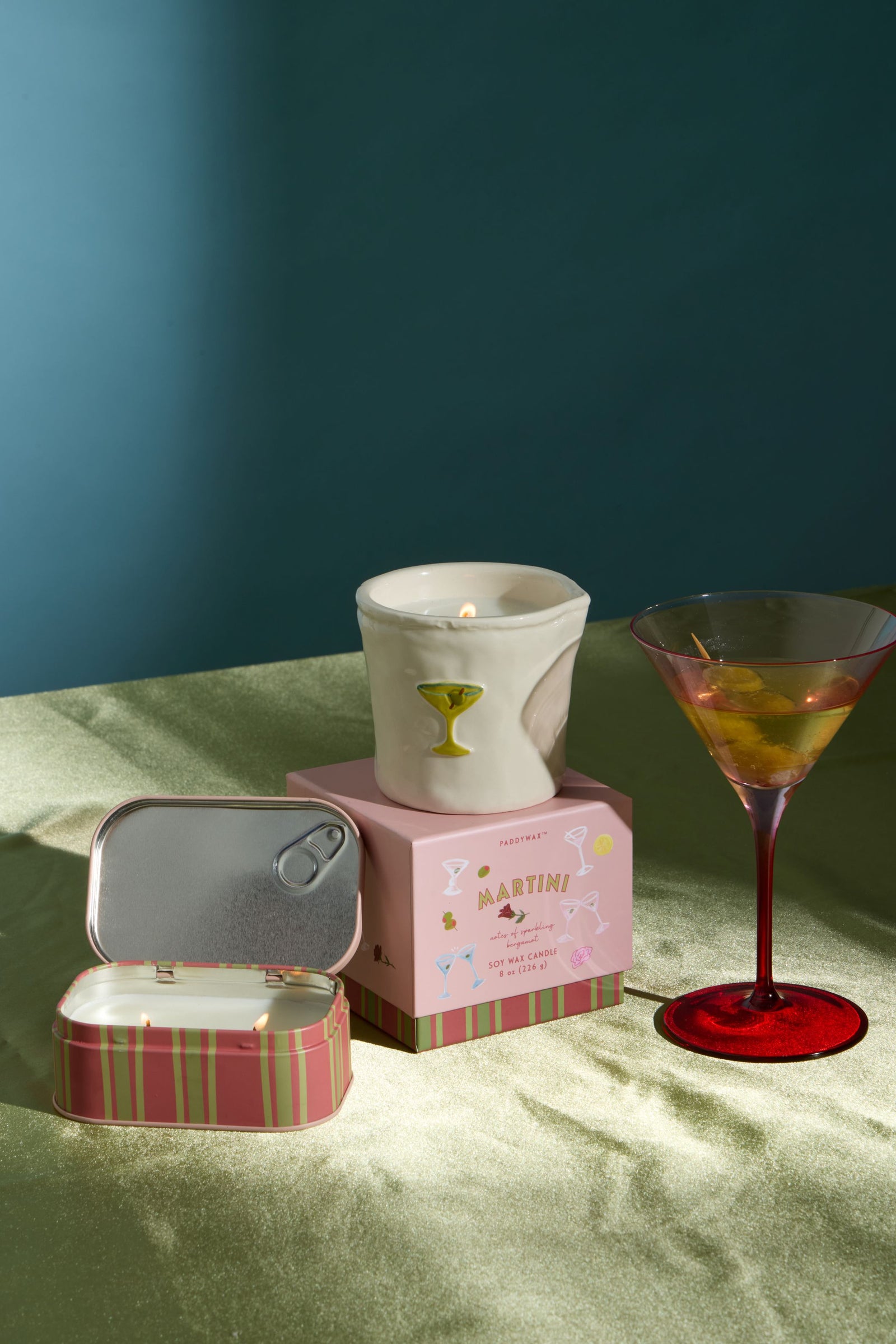 A styled product shot featuring a white ceramic cocktail glass candle holder with a lit candle, displayed on a pink patterned box labeled 'MARTINI'. In the foreground, an open pink striped tin with a silver pull-tab lid sits next to a red-stemmed martini glass filled with an olive.
