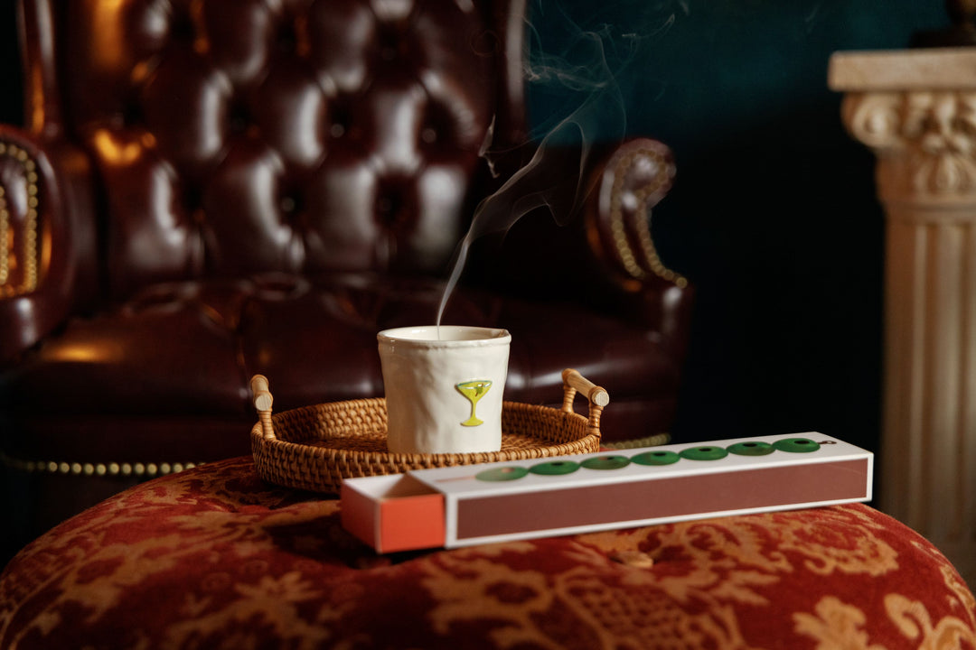 A white ceramic candle with martini motif sits on a woven tray, with smoke rising from it. In front of it is a matchbox-style container with green olive details, set on a red patterned velvet surface in front of a dark brown leather tufted armchair.